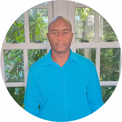 Man wearing a bright blue button-up shirt standing indoors in front of a window with green plants outside.