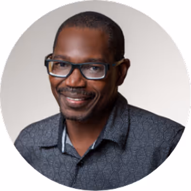 Smiling man wearing glasses and a patterned dark shirt against a light background.