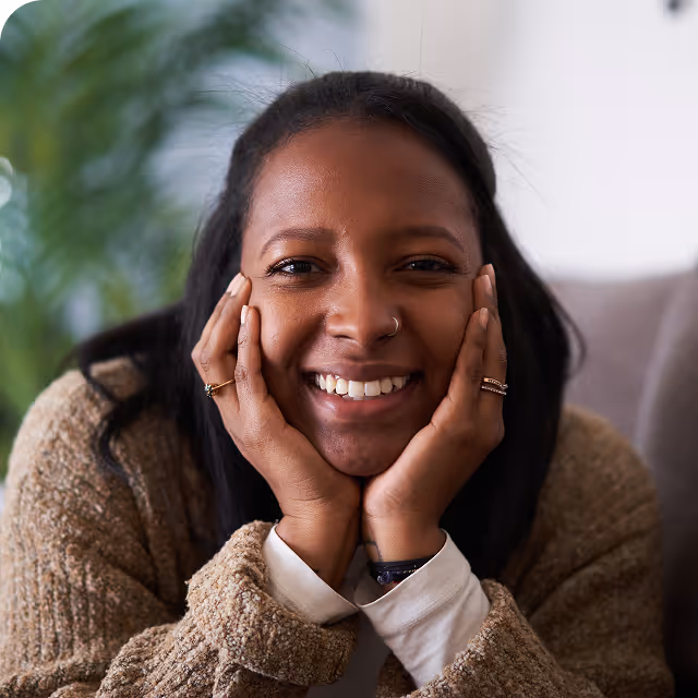 Smiling woman with dark hair resting her face in her hands, wearing a cozy beige sweater indoors.