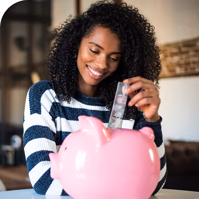 Smiling woman with curly hair inserting a dollar bill into a large pink piggy bank indoors.