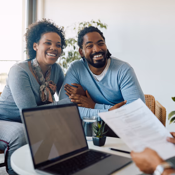 Smiling couple sitting together in a meeting, looking at a person holding a document across a table with a laptop.