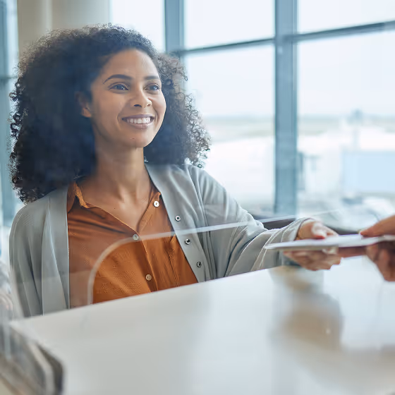 Smiling woman handing over a passport through an airport security plexiglass divider.